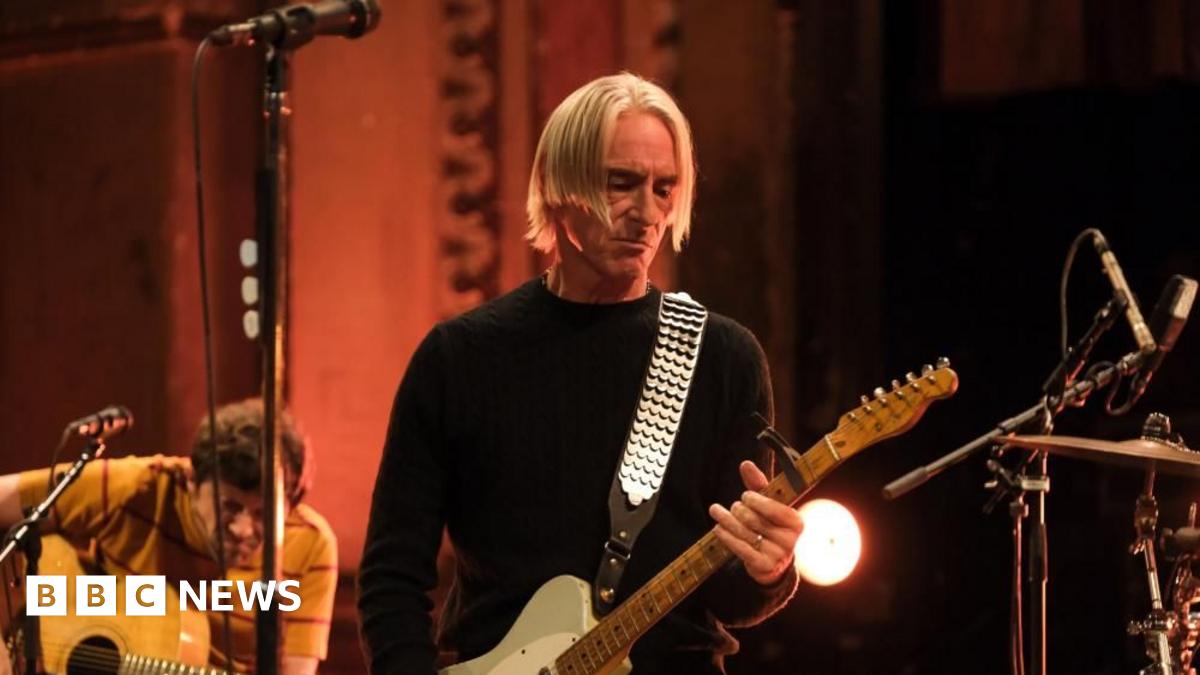 Paul Weller playing a guitar, with blond hair in a centre parting, looking down at the guitar. In the background is another guitarist and the edge of a drumkit.