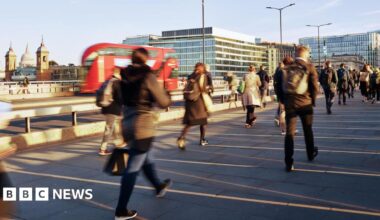 People walk across a bridge over the Thames in a blur as a red double decker bus drives past, with the sun low in the sky casting long shadows.
