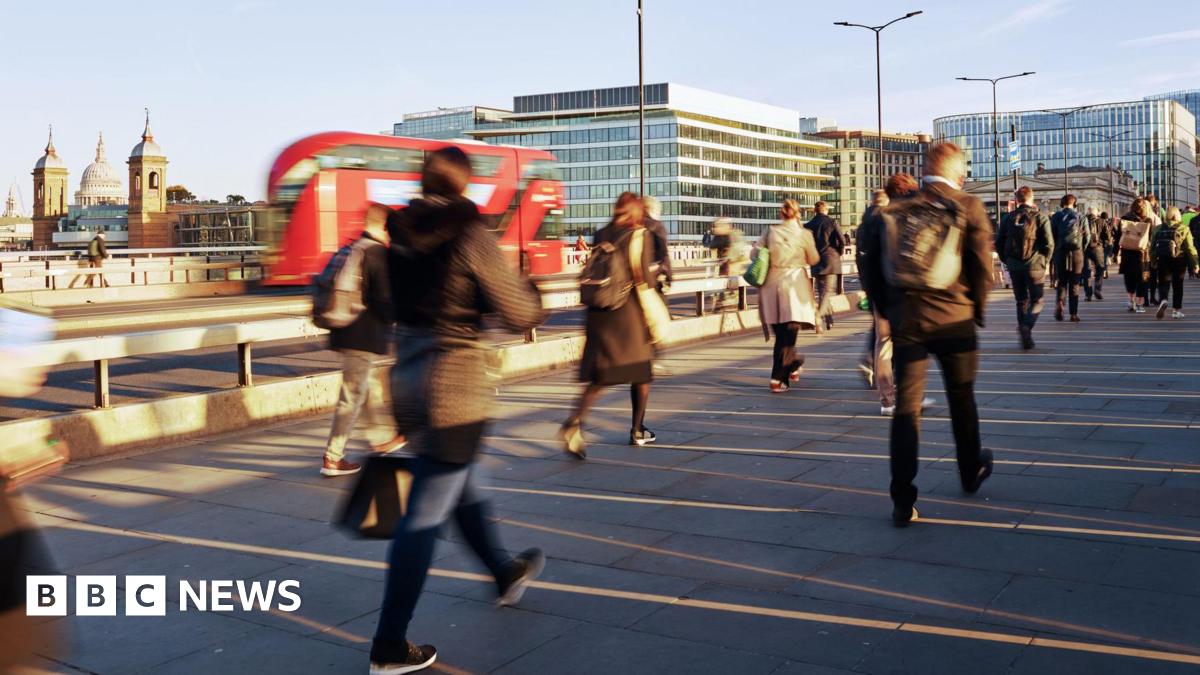 People walk across a bridge over the Thames in a blur as a red double decker bus drives past, with the sun low in the sky casting long shadows.