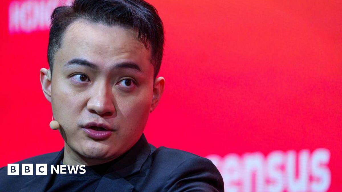 Justin Sun in black blazer and black T-shirt, with small beige microphone and ear piece in front of red background