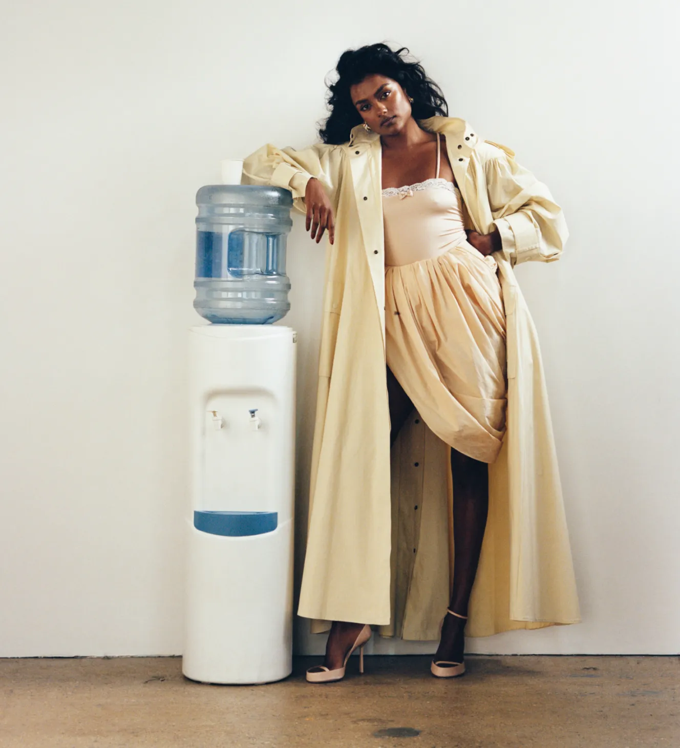 British actress Simone Ashley leans on a water cooler, wearing a long yellow coat over a pale yellow slip and heels.