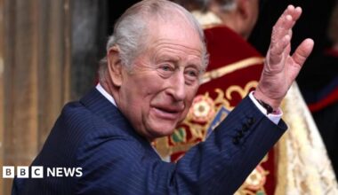 King Charles, dressed in a blue pinstriped suit, is smiling and waving at the entrance of St. George's Chapel, Windsor