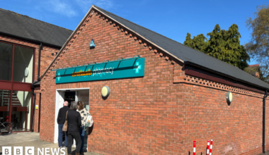 A brick building with the words Rowlands Pharmacy on a mainly green sign above a door. Three people are outside the door.