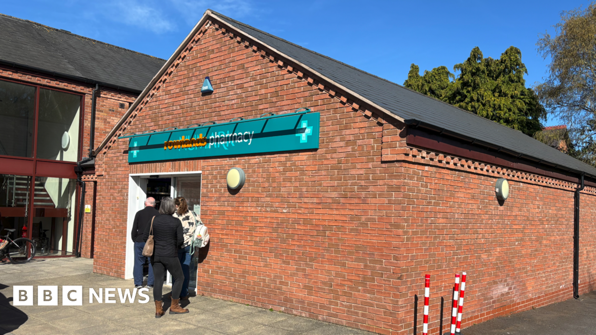 A brick building with the words Rowlands Pharmacy on a mainly green sign above a door. Three people are outside the door.