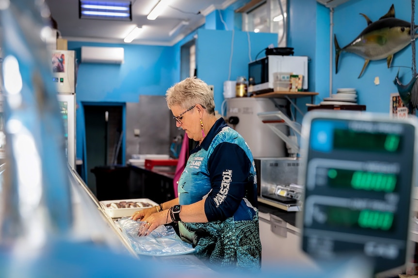 Woman with grey hair and glasses behind counter inside aqua coloured themed fish shop with scales in foreground