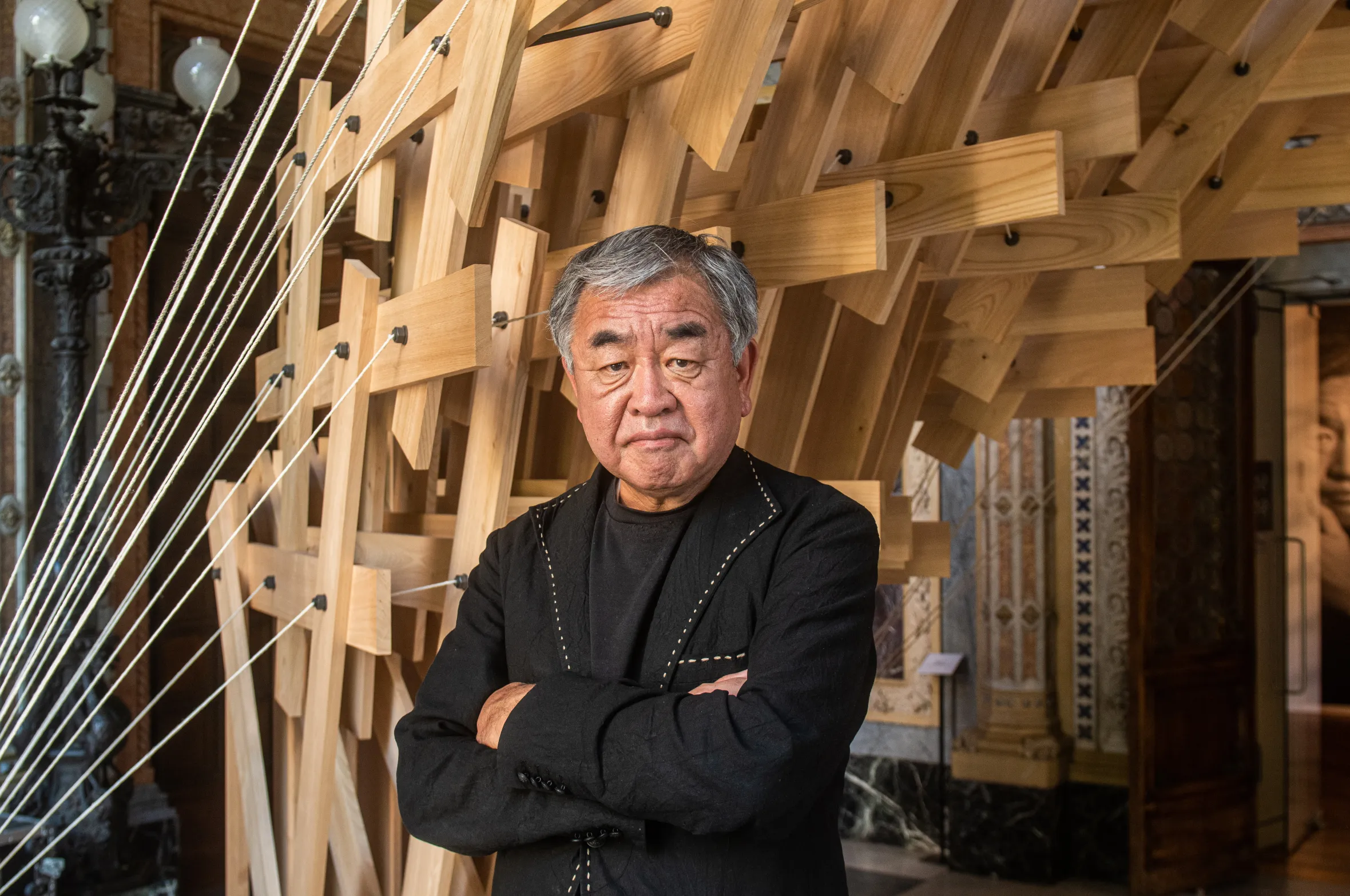 Architect Kengo Kuma stands with arms crossed in front of a wooden architectural installation.