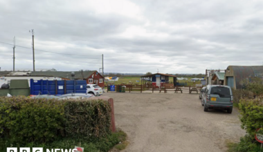 Multiple single storey buildings around a small yard. There are a few cars parked in the yard and some light aircraft can be seen on a grass airfield in the background.