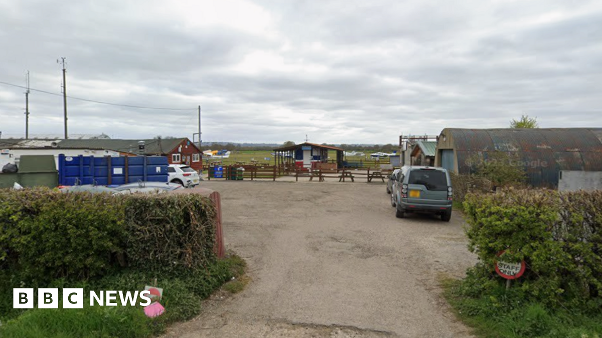 Multiple single storey buildings around a small yard. There are a few cars parked in the yard and some light aircraft can be seen on a grass airfield in the background.