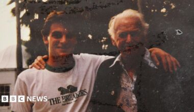 Nick Weston in a t-shirt and Douglas Fairbanks Jr in a floral shirt and jacket posing for a photograph in a garden. They have their arms around each other and are smiling.