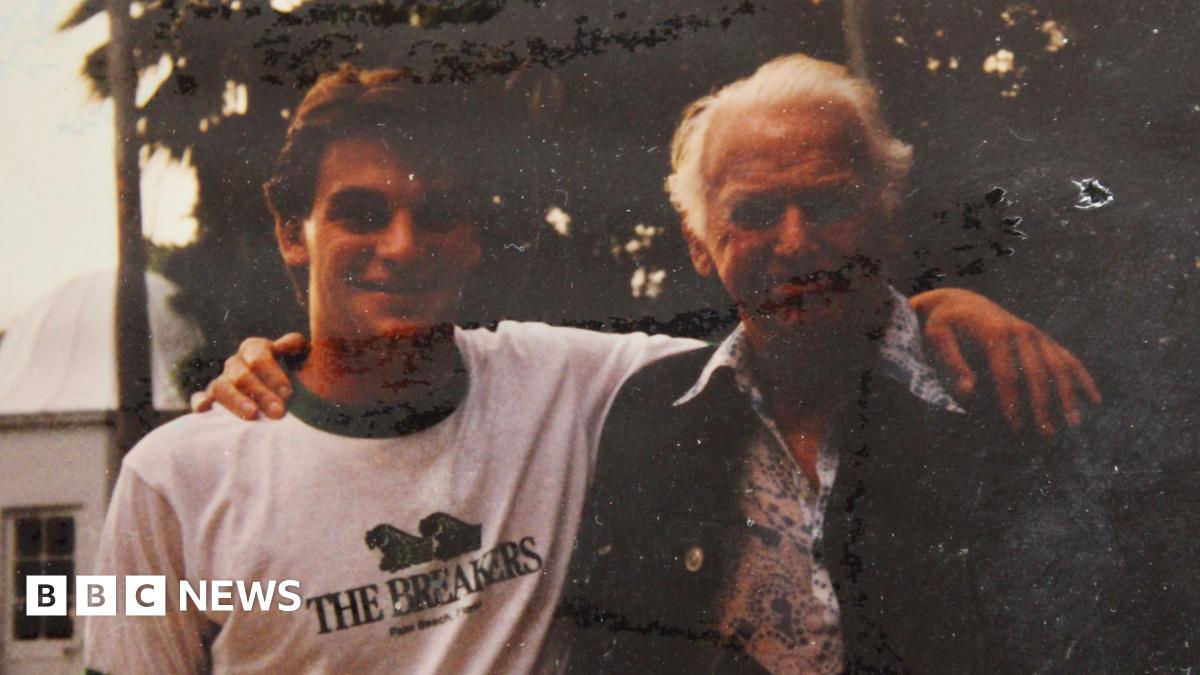 Nick Weston in a t-shirt and Douglas Fairbanks Jr in a floral shirt and jacket posing for a photograph in a garden. They have their arms around each other and are smiling.