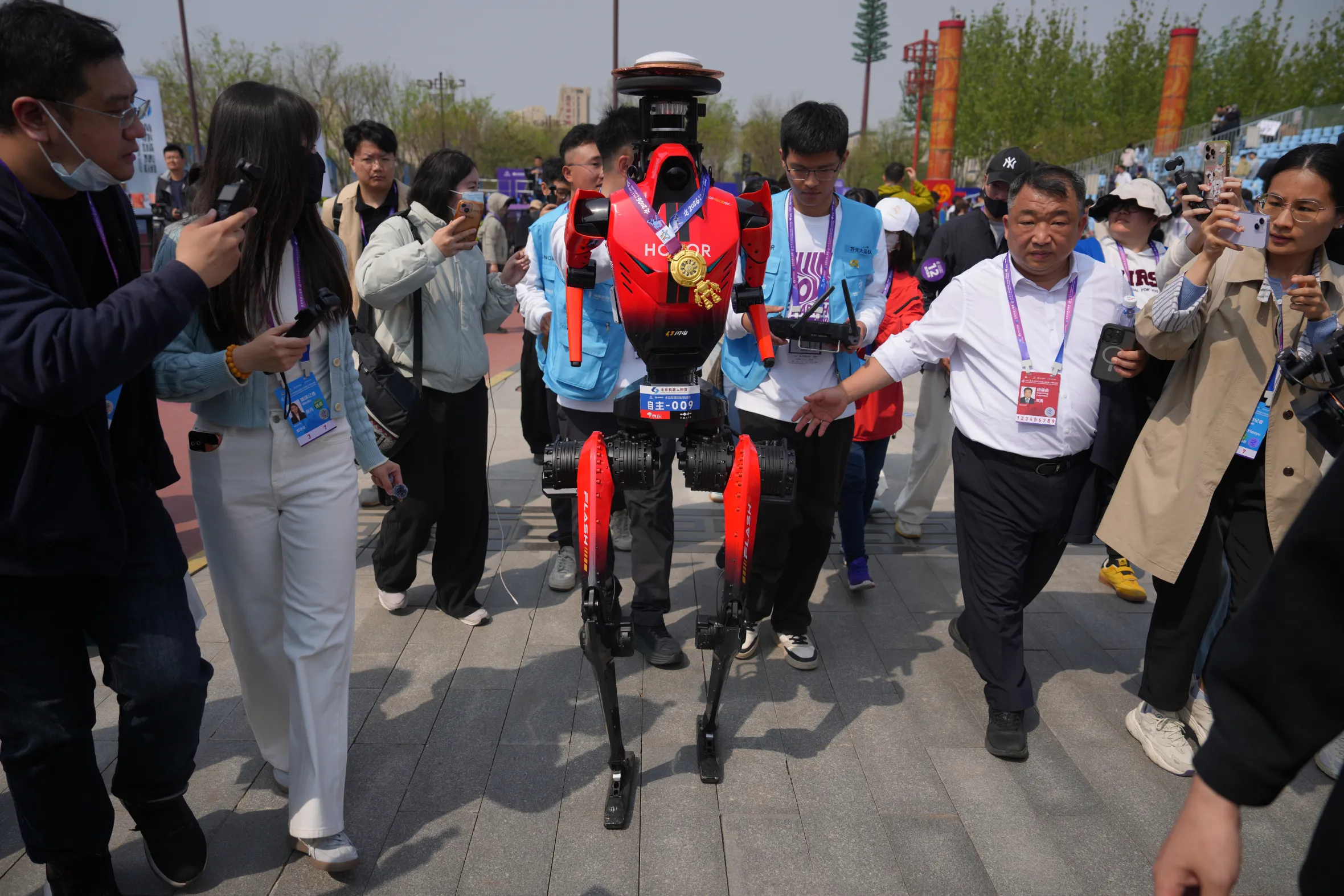 The winner of the humanoid robot half-marathon, a red and black robot with a medal around its neck, is surrounded by journalists.