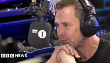 Scott Mills leaning into the microphone in the Radio 1 studio with headphones on.