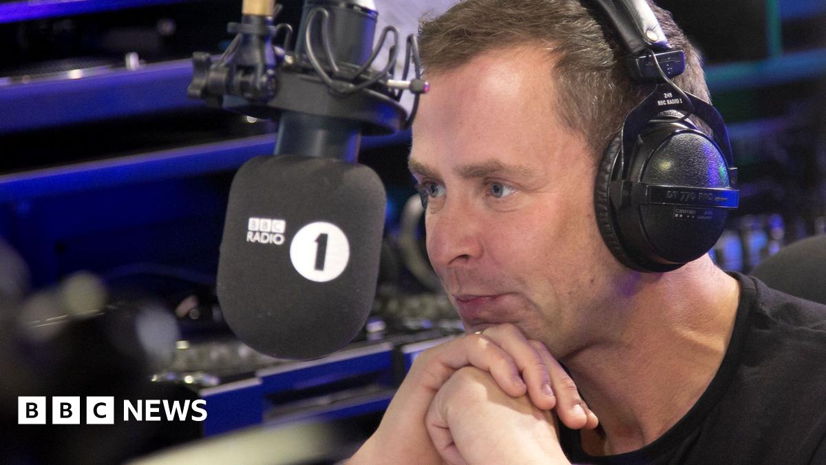 Scott Mills leaning into the microphone in the Radio 1 studio with headphones on.