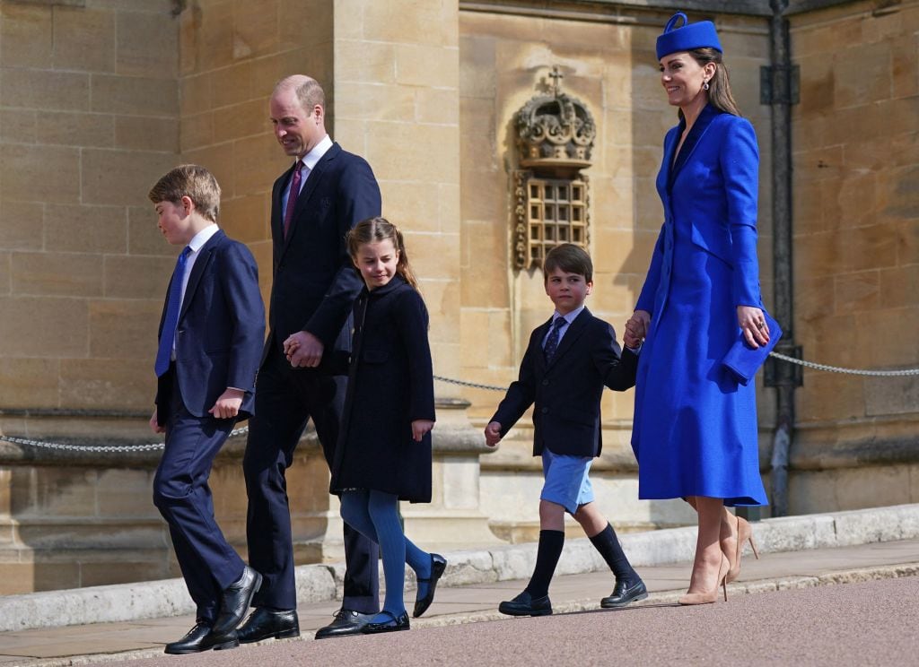 Two adults and three children walk beside a large golden stone building.