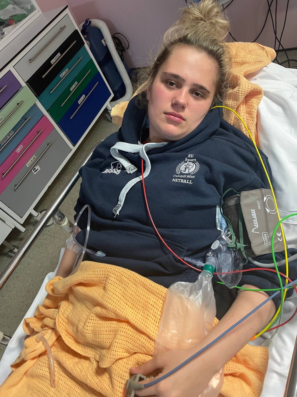 An individual, possibly a patient, reclines in a hospital bed, accompanied by various medical equipment. The person is dressed in a dark blue hoodie and has a medical device attached to their arm. Their expression is contemplative as they look directly at the camera, with a medical monitor visible in the background.