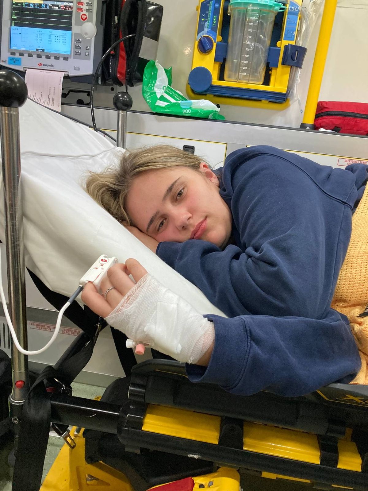 An individual is lying on a hospital bed, resting their head on a white pillow. They are wearing a blue long-sleeve shirt and have a white bandage on their left arm. The person appears to be holding a device connected to a wire.
