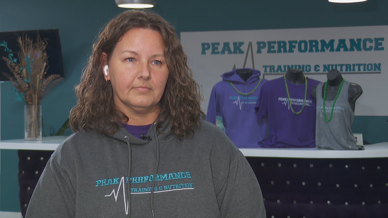 A woman stands in a sweater, looking off camera. Behind her there is a wall with the name of her business, 'Peak Performance Training & Nutrition'