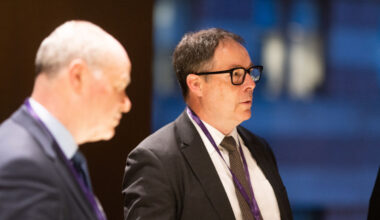 Ken Murphy delivering a keynote speech at a business conference, wearing a suit and gesturing at a presentation screen.