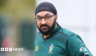 Monty Panesar has a black headscarf on and wears black glasses. He has black facial hair and is wearing a teal coloured t-shirt with a Twickenham Cricket Club logo on its breast. It is raining around him