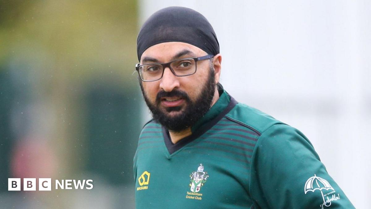 Monty Panesar has a black headscarf on and wears black glasses. He has black facial hair and is wearing a teal coloured t-shirt with a Twickenham Cricket Club logo on its breast. It is raining around him