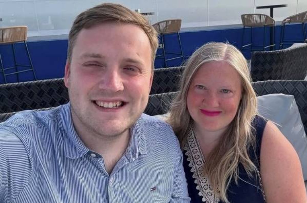 An image of an adult male and an adult female smiling and posing together, likely at an outdoor event. The background features what appears to be a blue-colored structure, possibly part of a venue or event space.