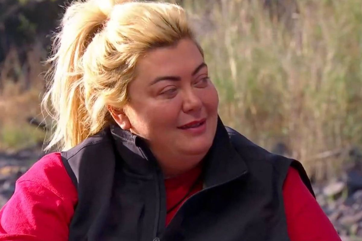 An adult female with blonde hair is standing outdoors in a forested area. She is wearing a red shirt and a black vest, with a smile on her face, looking to the side.