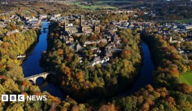 Aerial view of Durham in sunshine. The cathedral, castle and associated buildings stand in the bend of the river with the city spread out on either side behind. The scene is saturated in colour, with the river a deep blue, fringed by trees in a variety of autumnal colours.