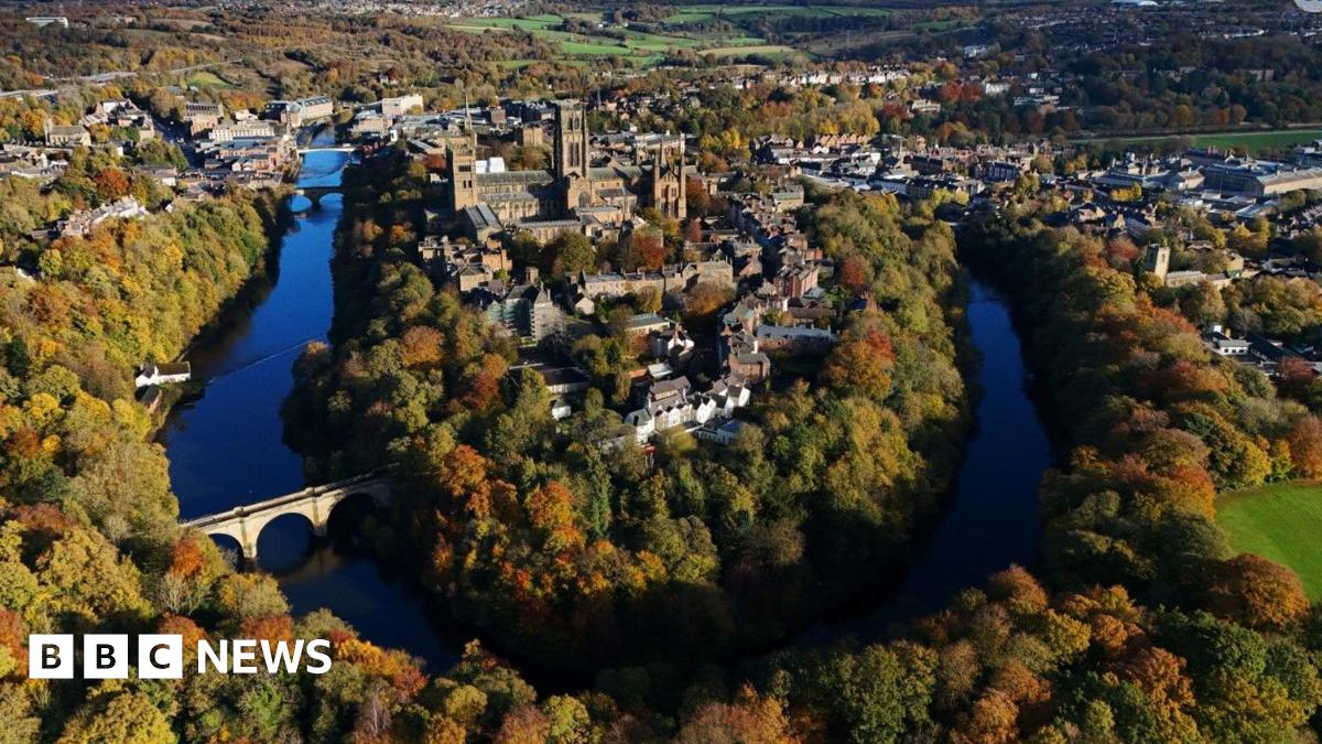 Aerial view of Durham in sunshine. The cathedral, castle and associated buildings stand in the bend of the river with the city spread out on either side behind. The scene is saturated in colour, with the river a deep blue, fringed by trees in a variety of autumnal colours.
