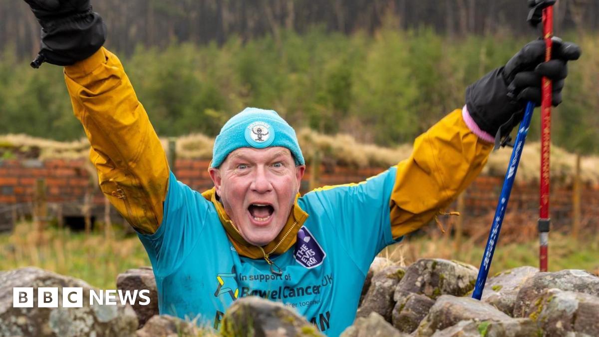 A man at the bottom of Pen y Fan. There are rocks around him. The man has a blue t-shirt and under a yellow long sleeved shirt with a blue hat, black sunglasses and black shorts. His arms are outstretched like his is cheering.