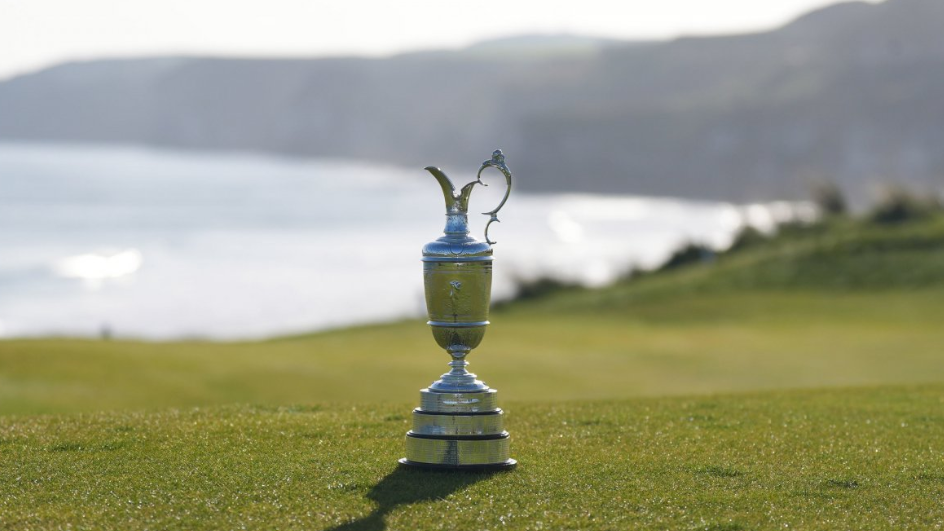 The silver claret jug sits on grass at Royal Portush Golf Club. You can see the headland and the sea in the background.