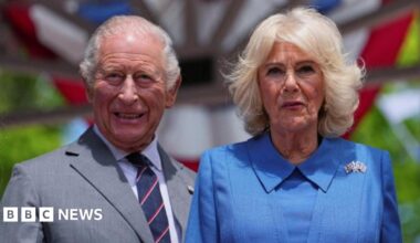 King Charles and Queen Camilla look directly ahead in the sunshine during their US state visit. He is dressed in a grey suit, with shirt and tie. Camilla is wearing a blue outfit with a brooch featuring the US and British flags.