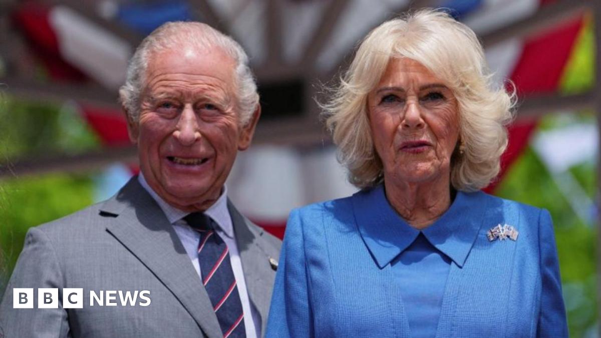 King Charles and Queen Camilla look directly ahead in the sunshine during their US state visit. He is dressed in a grey suit, with shirt and tie. Camilla is wearing a blue outfit with a brooch featuring the US and British flags.