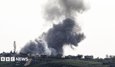 A large grey bundle of smoke rising from a village on a hillside. The smoke cloud fills most of the picture and is dramatic against the backdrop