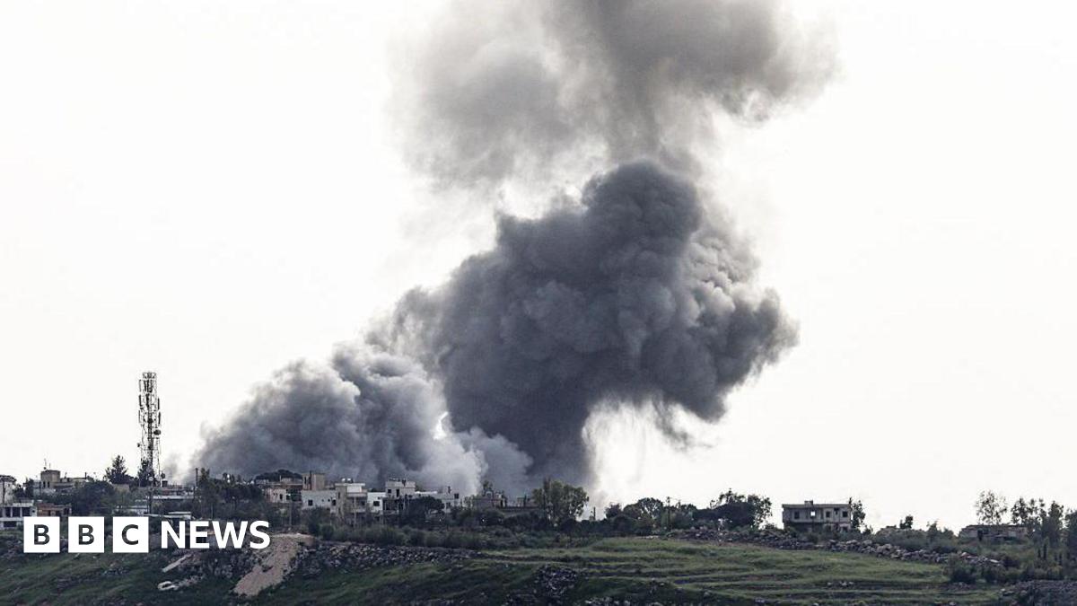 A large grey bundle of smoke rising from a village on a hillside. The smoke cloud fills most of the picture and is dramatic against the backdrop