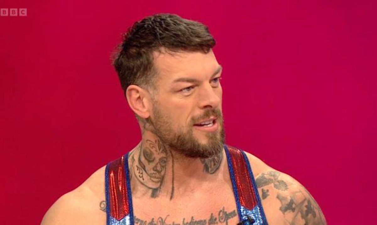 An individual with visible tattoos on their neck and arms stands against a red background, appearing to speak or participate in an event.
