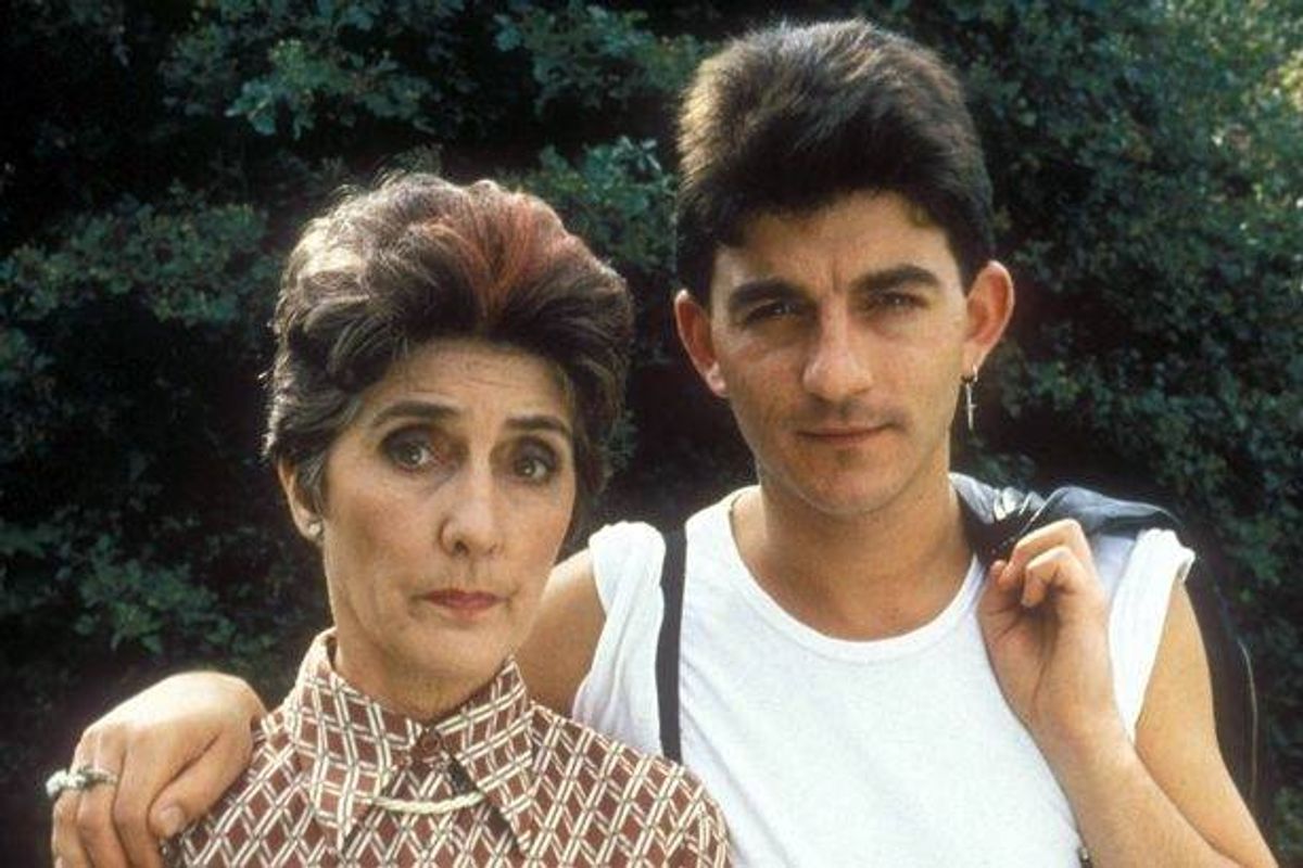 An adult female and a young male are standing side by side, posing for a photograph in an outdoor setting, with lush greenery forming a backdrop behind them.