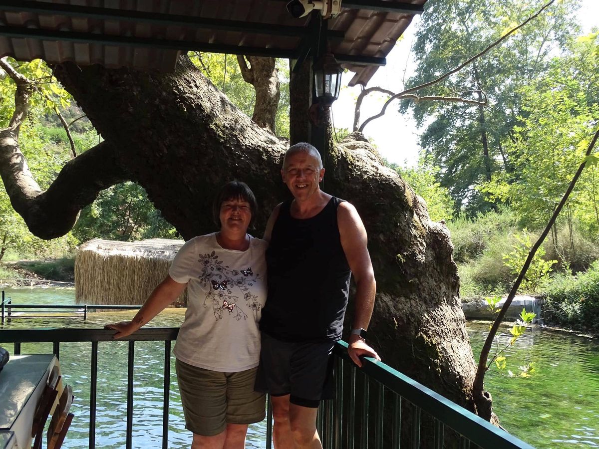 Two individuals are standing beside a massive tree on a wooden deck, with lush greenery and a tranquil pond in the background.