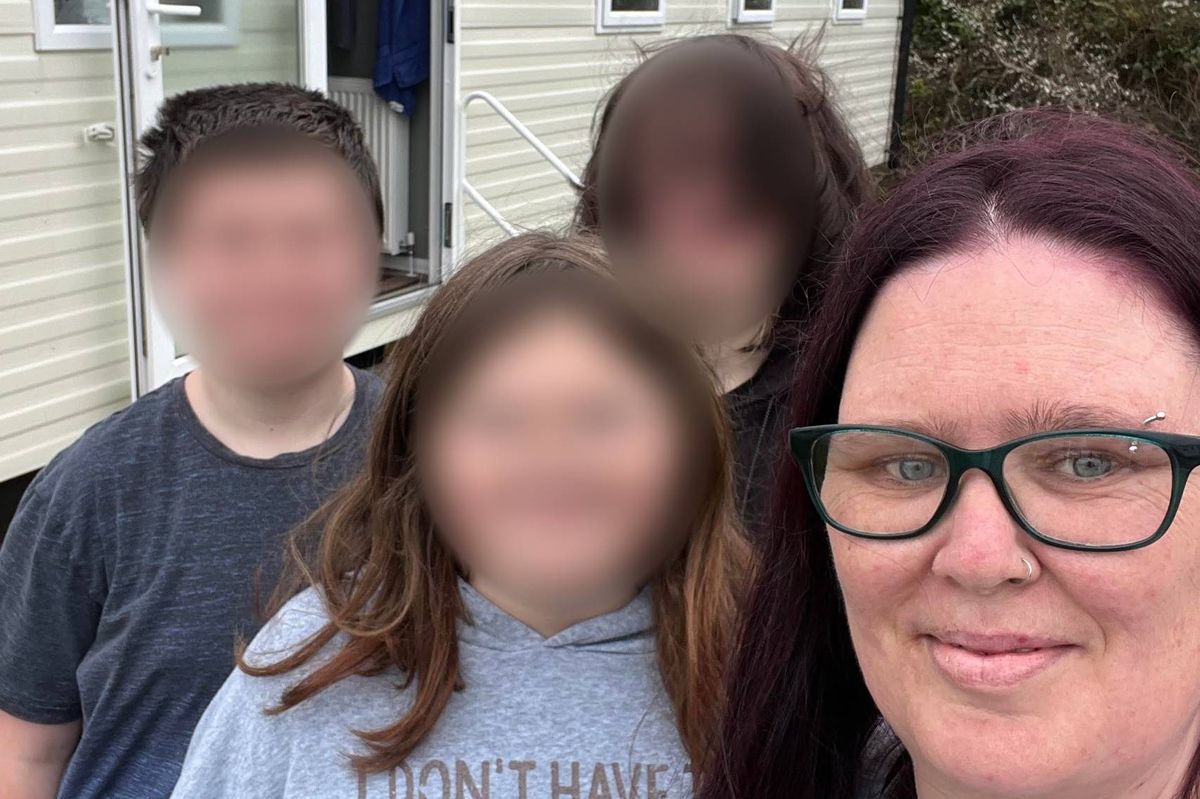 A woman with glasses smiles in front of a house, with several individuals standing behind her, their faces blurred.