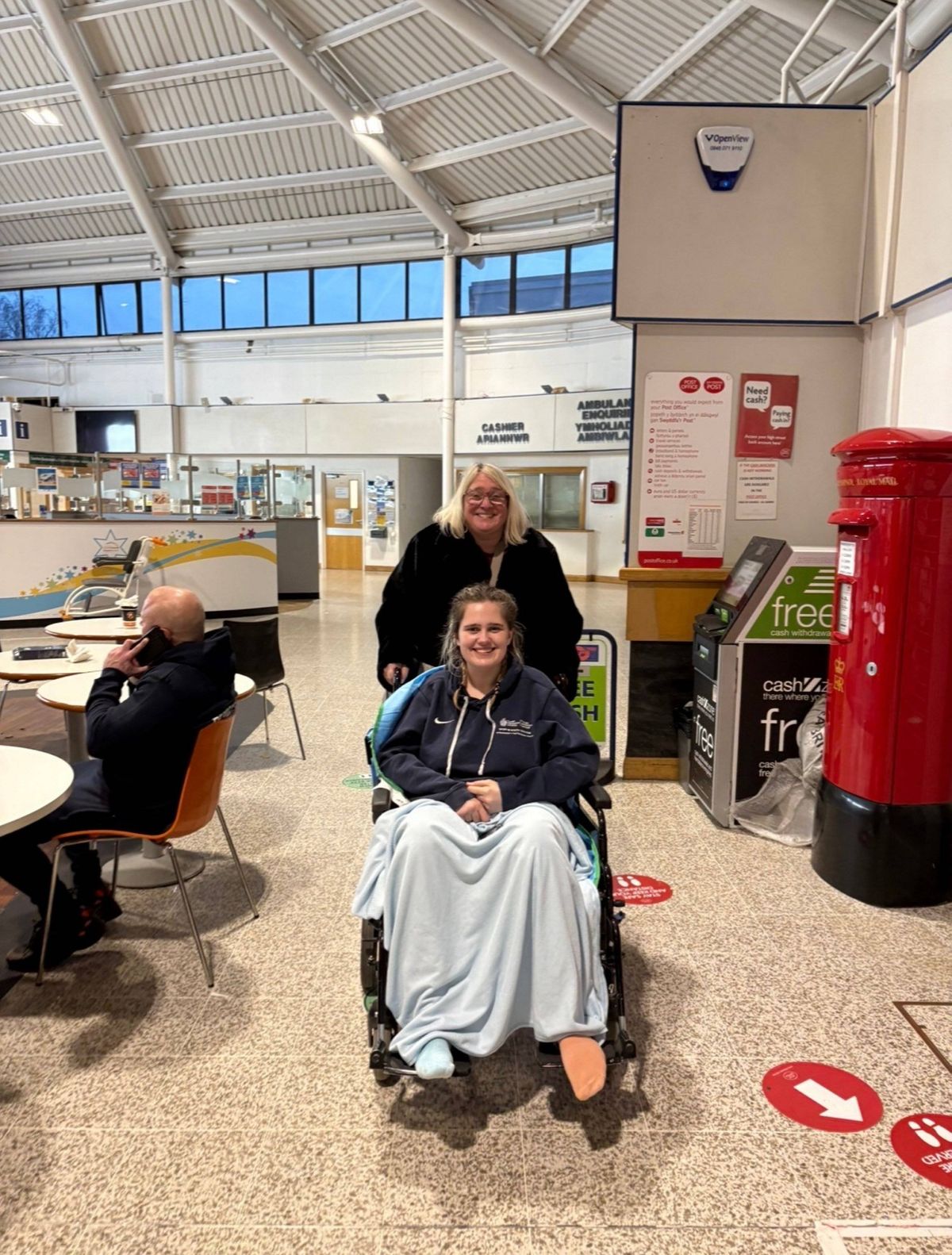 An indoor area with people seated at tables, one individual in a wheelchair accompanied by another person standing behind them, and various objects such as a red post box and a circular sign placed on the floor.