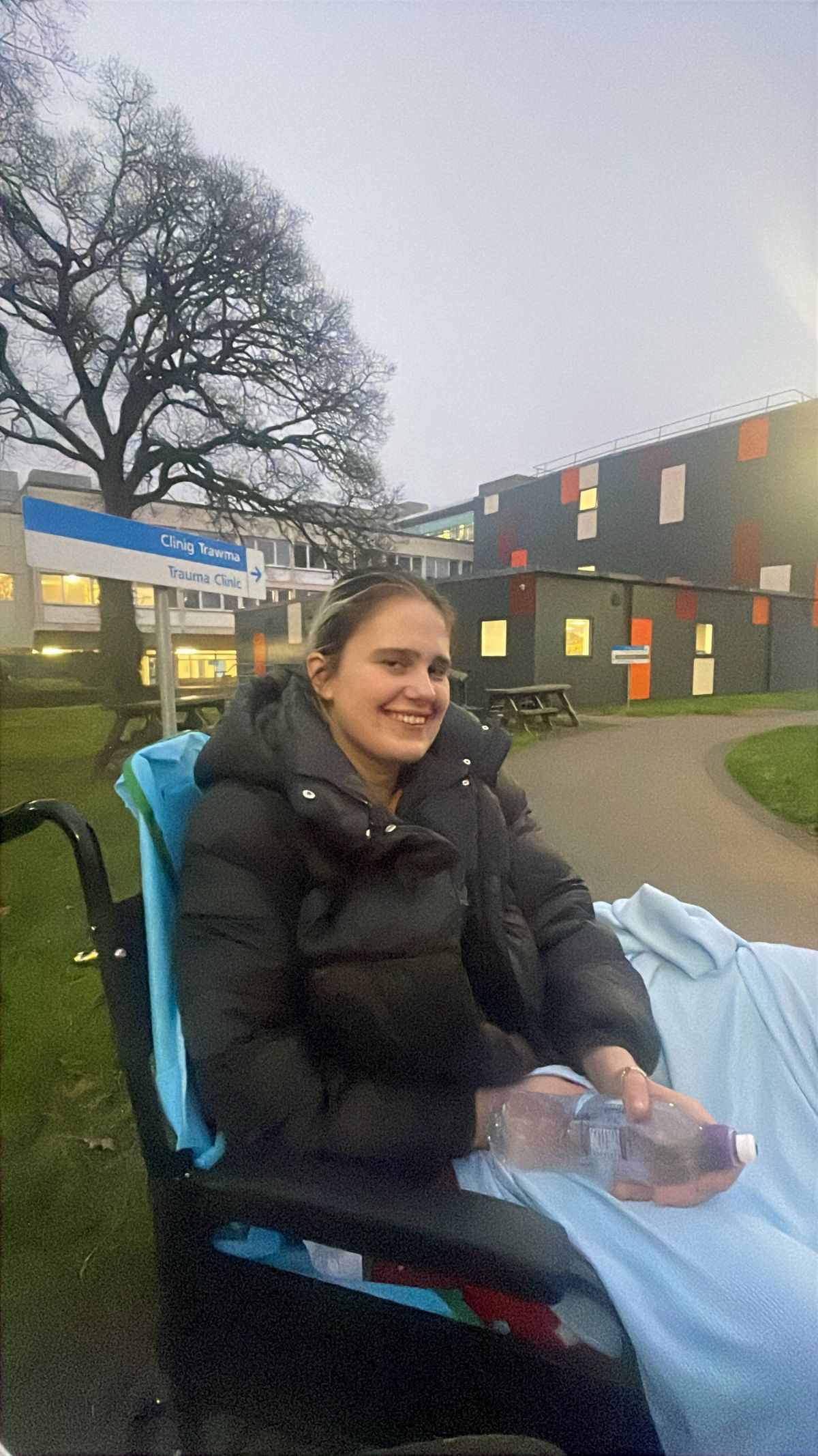 An individual wearing a black coat sits in a wheelchair outside of a building, with a tree and pathway in the background