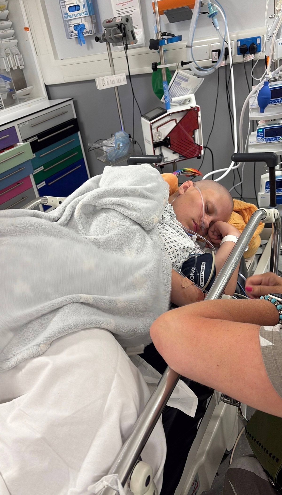 An individual is resting on a hospital bed, covered with a white blanket, while various medical devices and equipment surround the bed, indicating a hospital setting