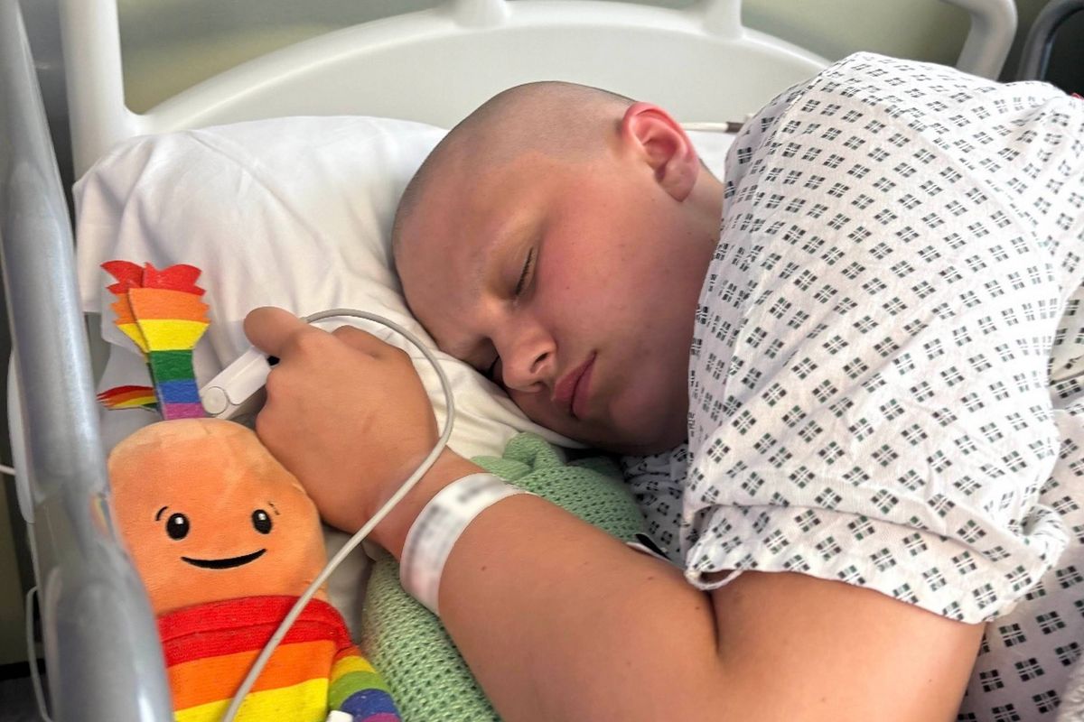 An individual resting in a hospital bed, accompanied by a stuffed toy, lies on their side with an arm resting on the pillow, dressed in a hospital gown, and connected to medical equipment