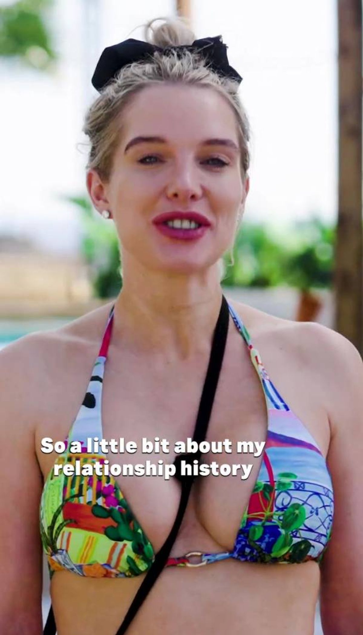 A close-up portrait of an individual with short blonde hair, wearing a sleeveless top, standing in an outdoor setting. The individual has a slight smile and appears to be engaged in a conversation. The background features blurred greenery, suggesting a natural environment.