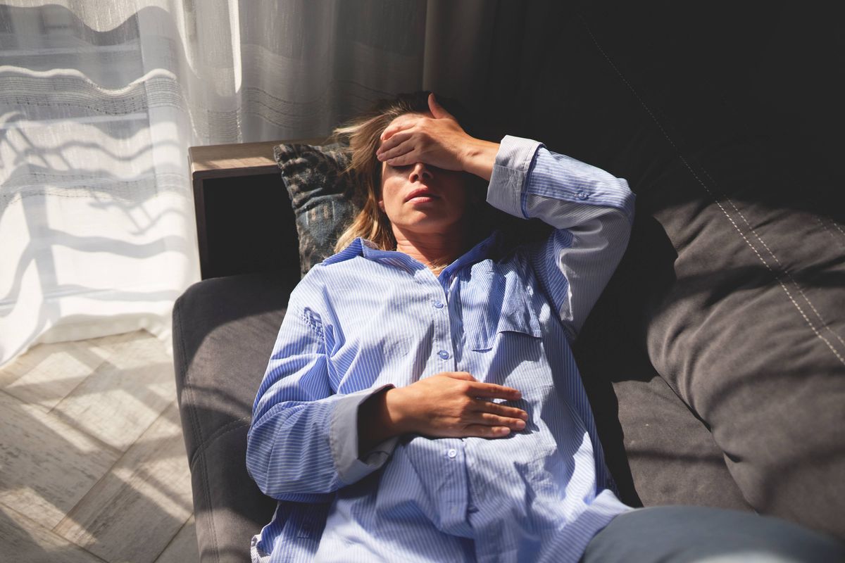 A woman lying on a couch with her hand on her forehead and eyes closed, indicating exhaustion or stress. The indoor scene is brightly lit by natural light coming through the window with curtains.