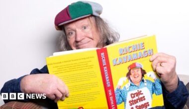 A man with long grey hair holding a yellow and red book, he is smiling at the camera.