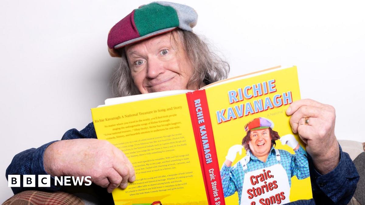A man with long grey hair holding a yellow and red book, he is smiling at the camera.
