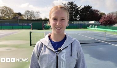 Daniella Britton wearing a grey and black hoodie and a blue t-shirt. She is stood on a blue and green tennis court and is staring at the camera smiling.