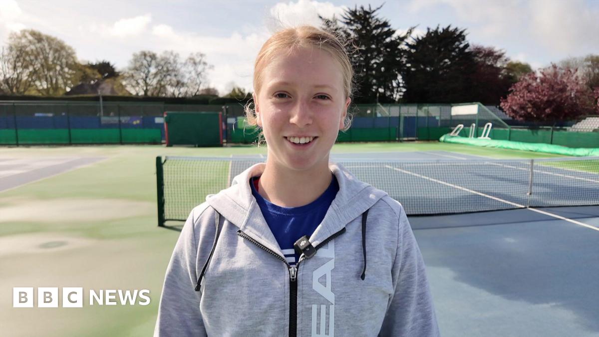 Daniella Britton wearing a grey and black hoodie and a blue t-shirt. She is stood on a blue and green tennis court and is staring at the camera smiling.