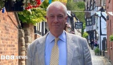 Ewen Sinclair is standing in a street in Ledbury, wearing a suit and tie - behind him are several black and white buildings.