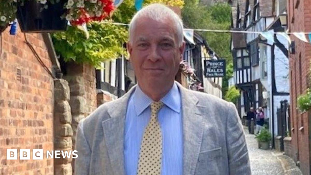 Ewen Sinclair is standing in a street in Ledbury, wearing a suit and tie - behind him are several black and white buildings.